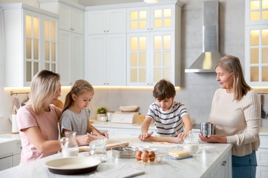 Photo of Little kids with their mother and grandmother making dough at white marble table in kitchen