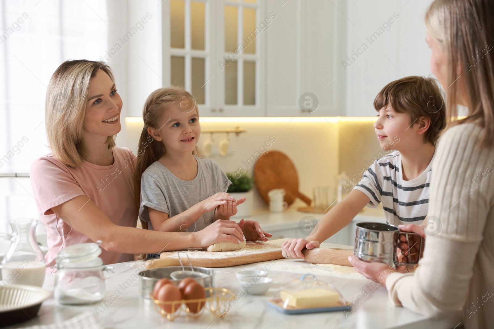 Photo of Little kids with their mother and grandmother making dough at white marble table in kitchen