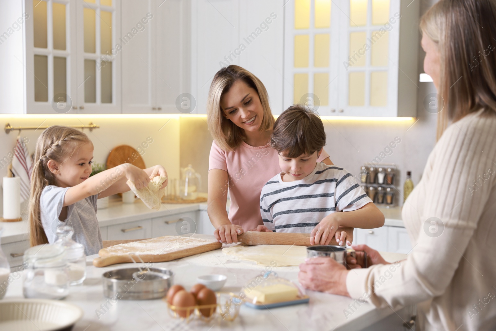 Photo of Little kids with their mother and grandmother making dough at white marble table in kitchen