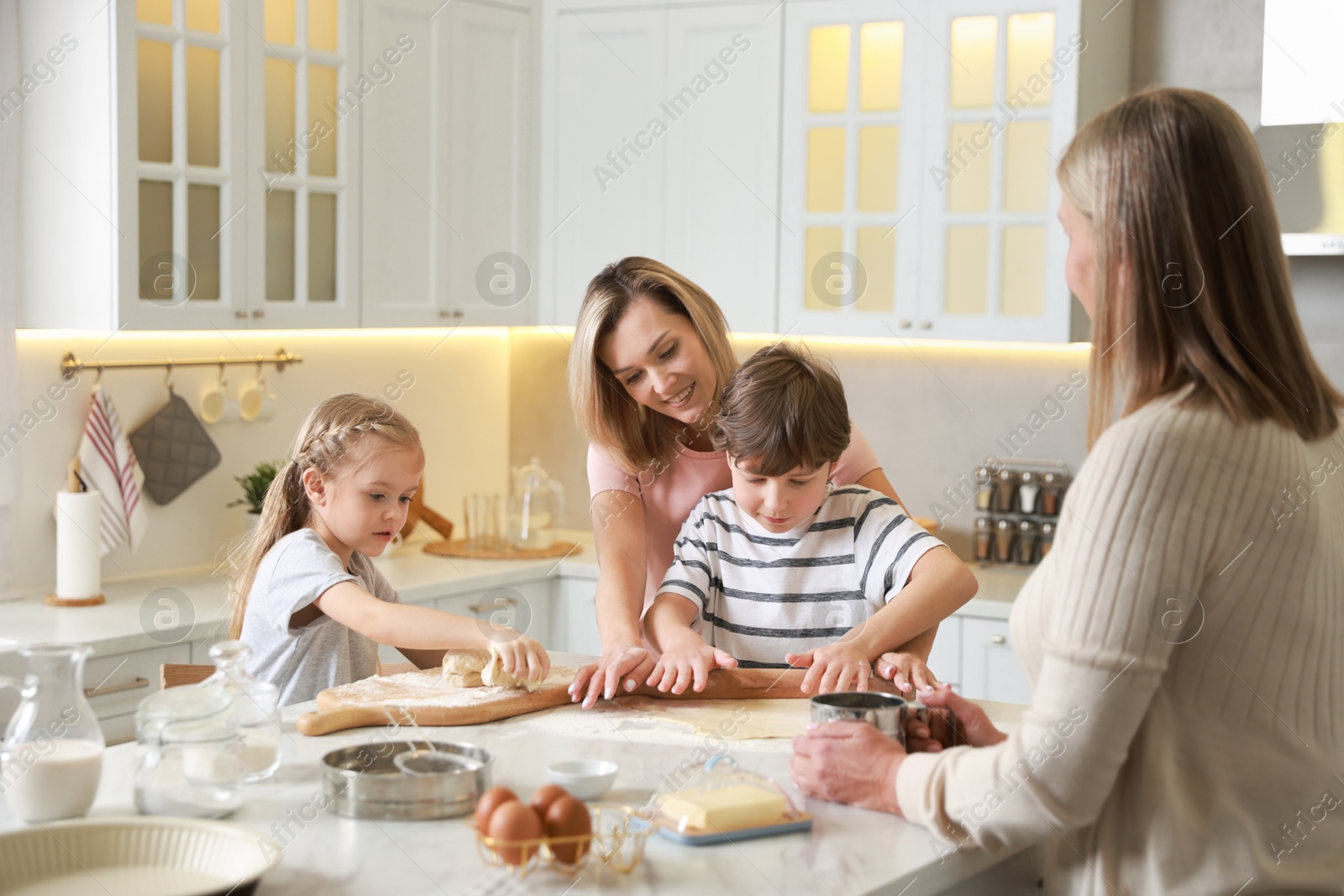 Photo of Little kids with their mother and grandmother making dough at white marble table in kitchen