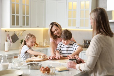 Photo of Little kids with their mother and grandmother making dough at white marble table in kitchen