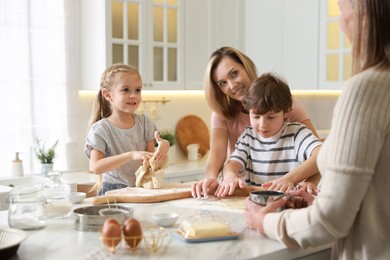 Photo of Little kids with their mother and grandmother making dough at white marble table in kitchen