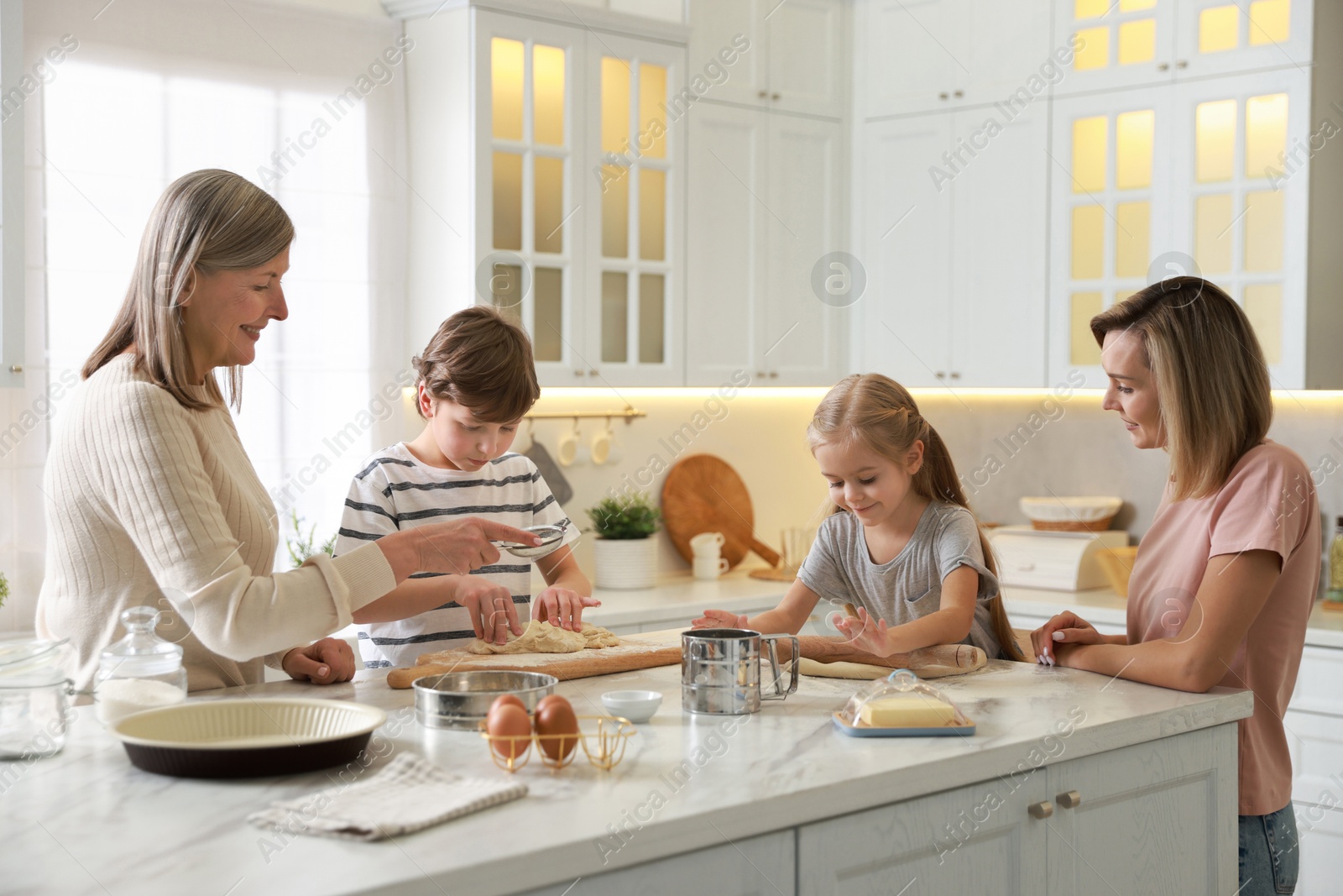 Photo of Little kids with their mother and grandmother making dough at white marble table in kitchen