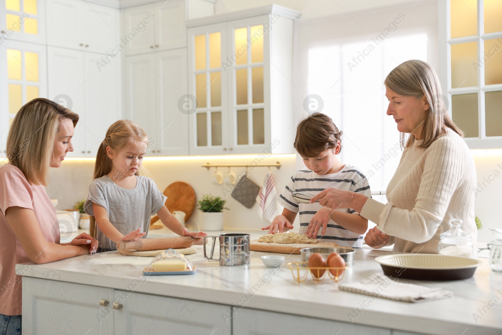 Photo of Little kids with their mother and grandmother making dough at white marble table in kitchen