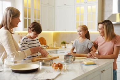 Photo of Little kids with their mother and grandmother making dough at white marble table in kitchen