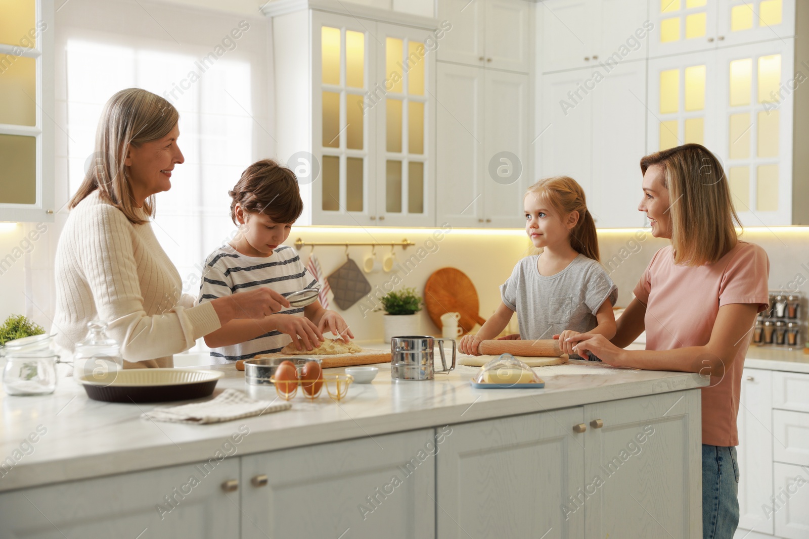 Photo of Little kids with their mother and grandmother making dough at white marble table in kitchen