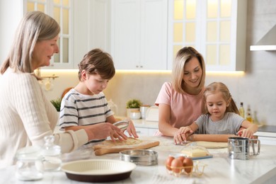 Photo of Little kids with their mother and grandmother making dough at white marble table in kitchen