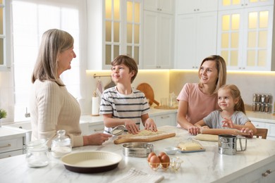 Photo of Little kids with their mother and grandmother making dough at white marble table in kitchen