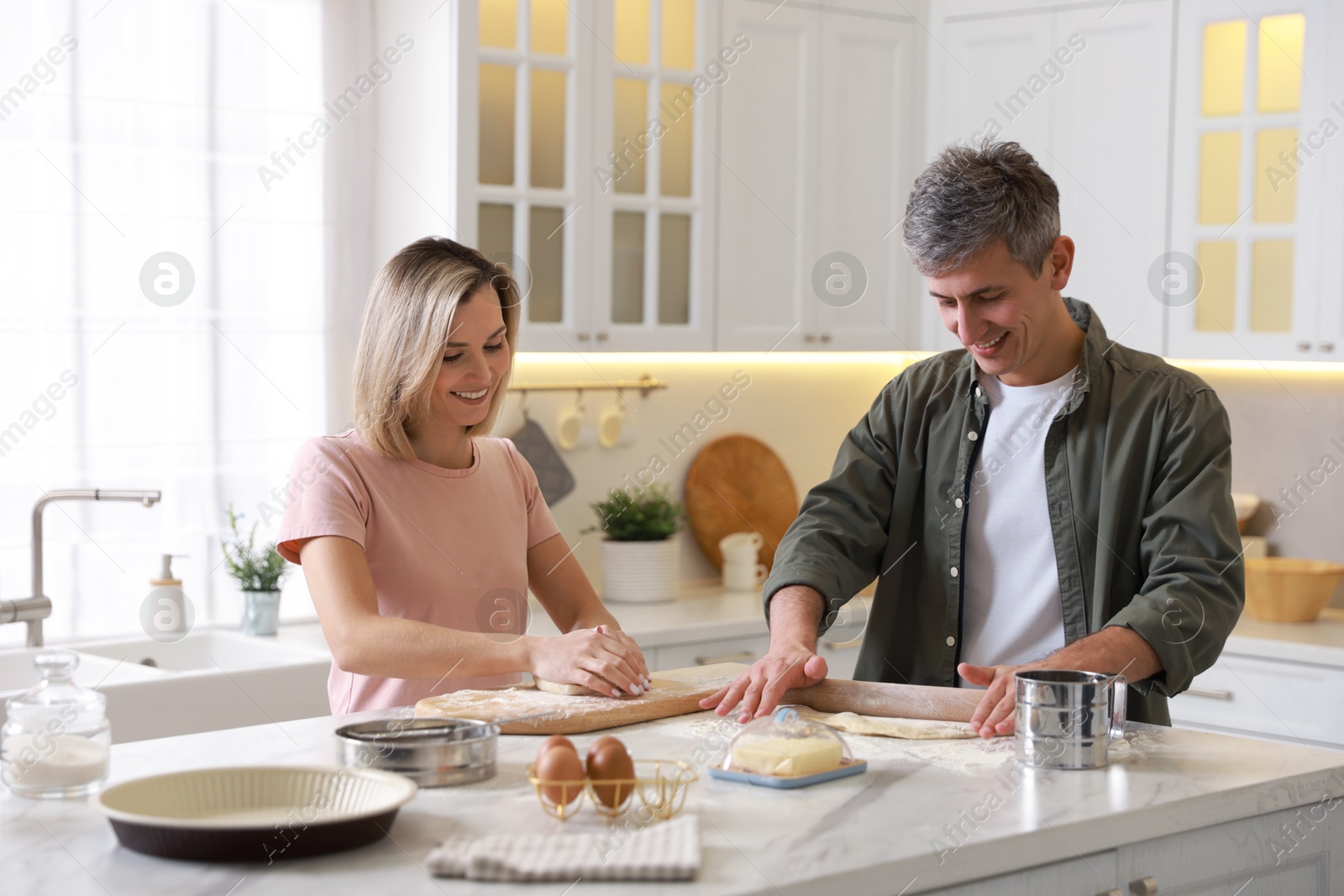 Photo of Happy couple kneading dough at white marble table in kitchen