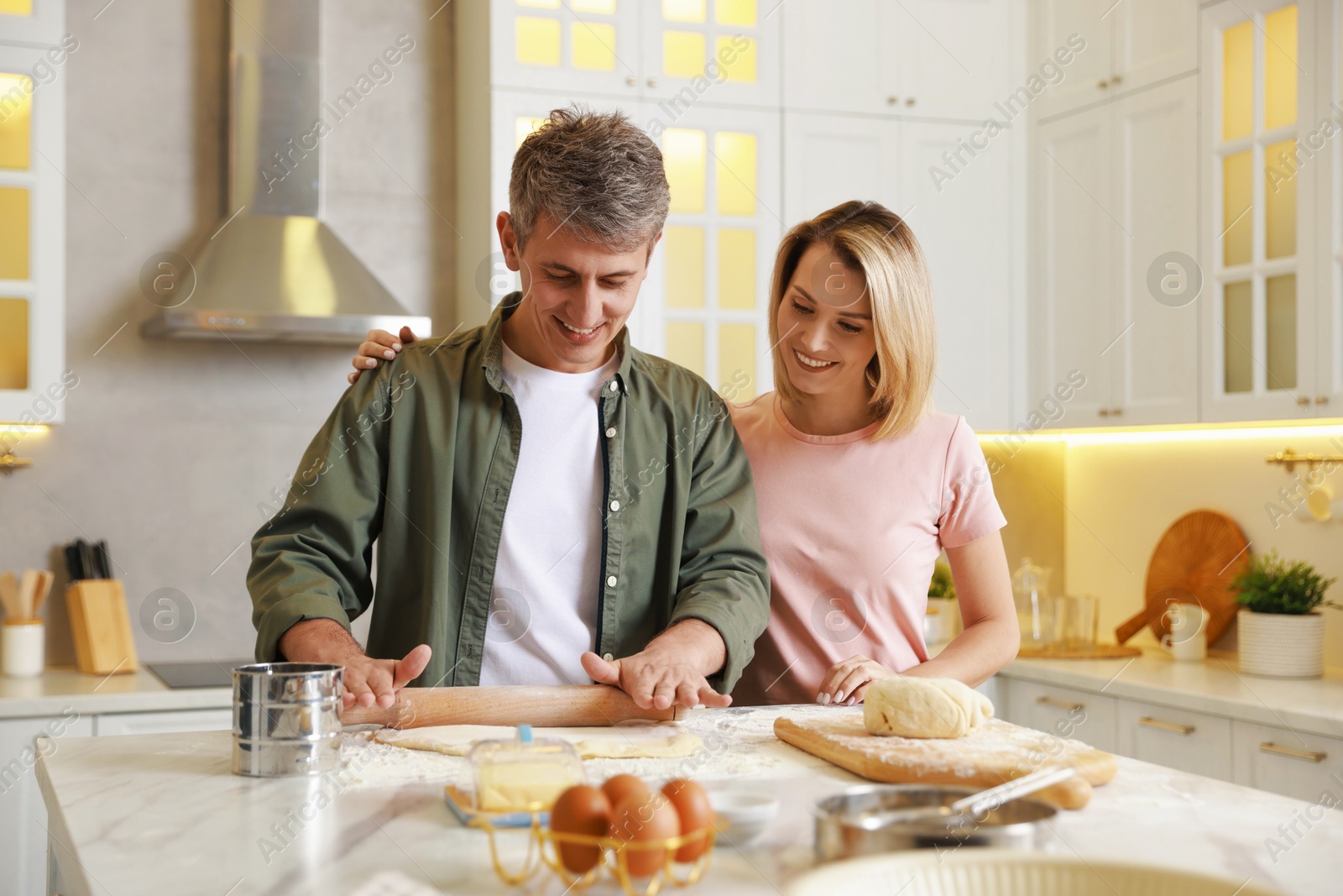 Photo of Happy couple shaping dough with rolling pin at white marble table in kitchen