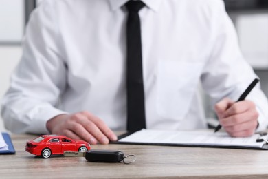 Photo of Man signing purchase agreement at wooden table, focus on key and car model. Buying auto