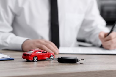 Photo of Man signing purchase agreement at wooden table, focus on key and car model. Buying auto