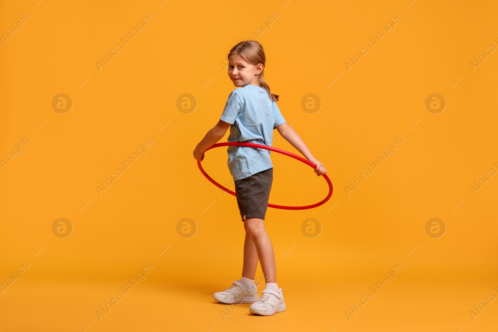 Photo of Cute little girl exercising with hula hoop on orange background, space for text