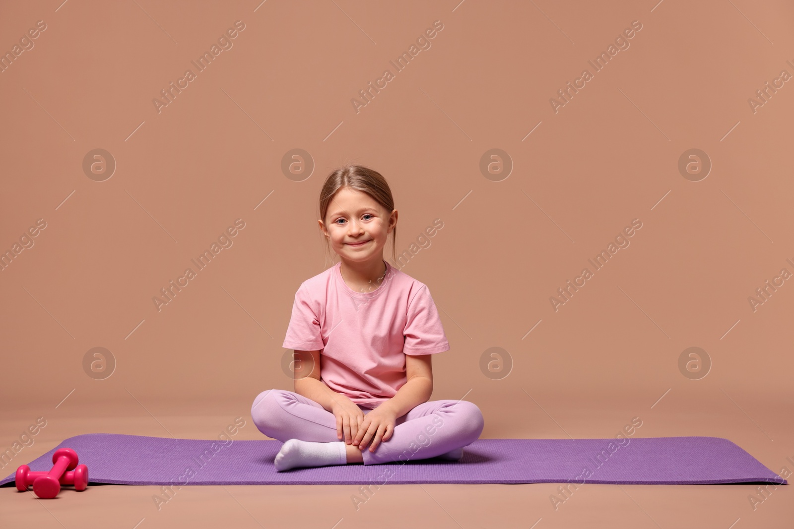 Photo of Cute little girl in sportswear sitting on fitness mat against dark beige background