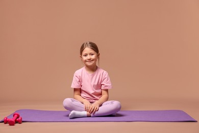 Photo of Cute little girl in sportswear sitting on fitness mat against dark beige background