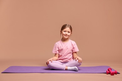 Photo of Cute little girl in sportswear sitting on fitness mat against dark beige background