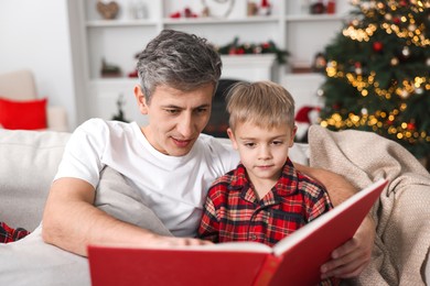 Photo of Father and son reading book together on sofa at home. Christmas holidays