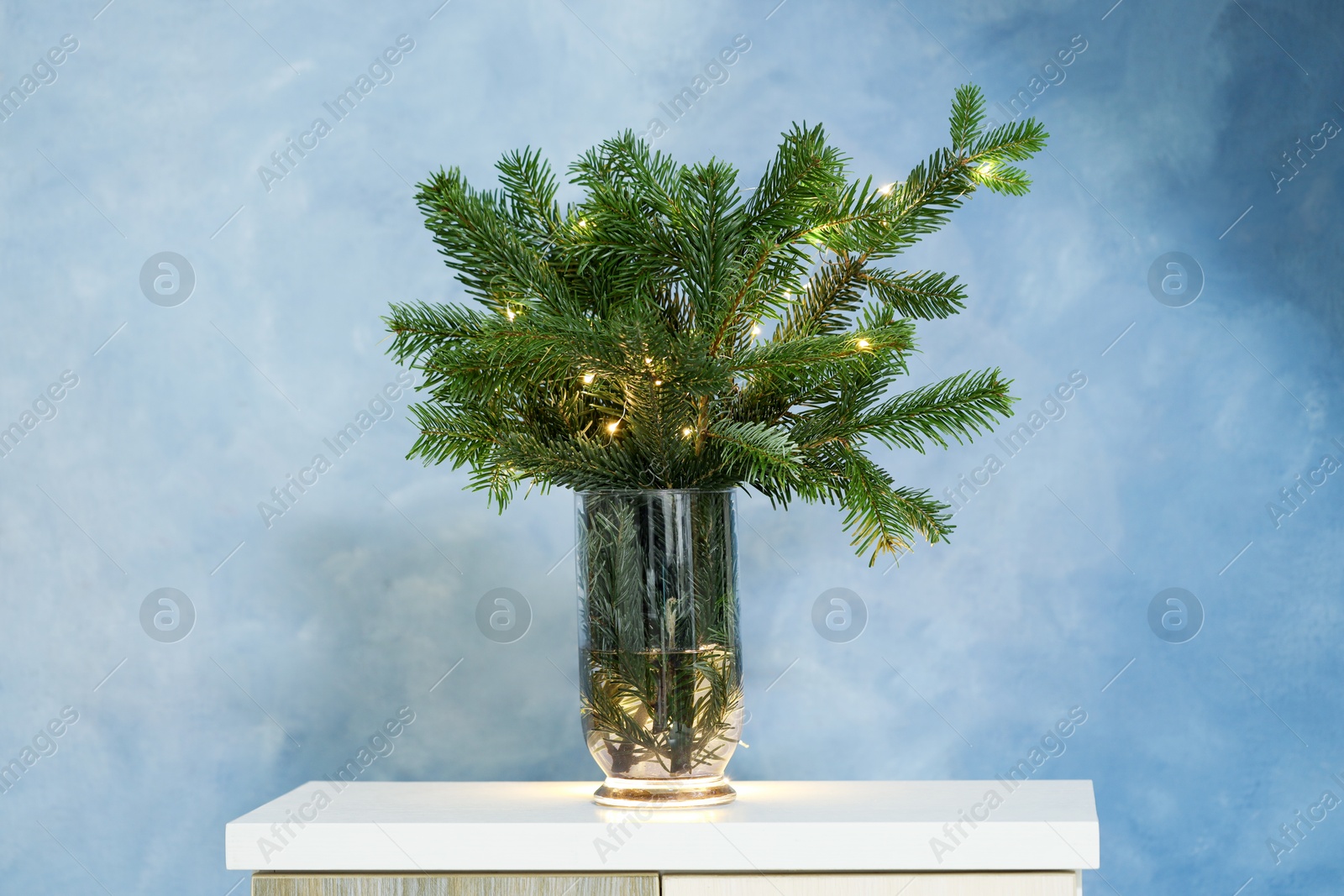 Photo of Beautiful fir tree branches decorated with Christmas lights on table near blue wall