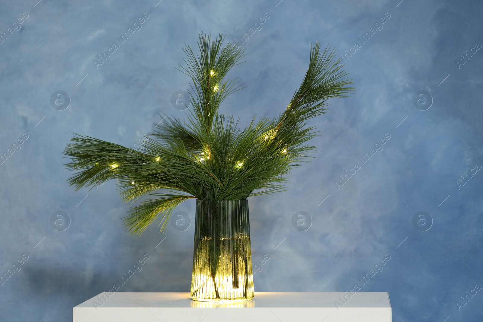 Photo of Beautiful pine tree branches decorated with Christmas lights on table near blue wall