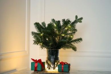 Photo of Beautiful fir tree branches decorated with festive lights and Christmas gifts on table near white wall