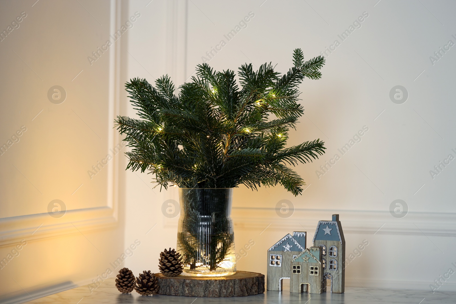 Photo of Beautiful fir tree branches decorated with Christmas lights on table near white wall