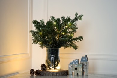 Photo of Beautiful fir tree branches decorated with Christmas lights on table near white wall