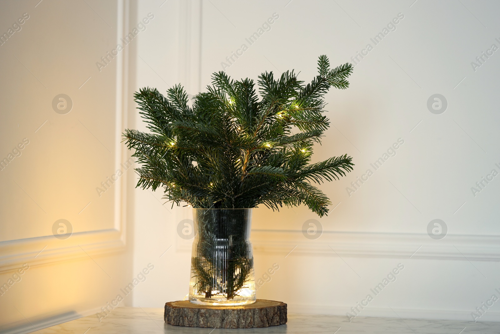 Photo of Beautiful fir tree branches decorated with Christmas lights on table near white wall