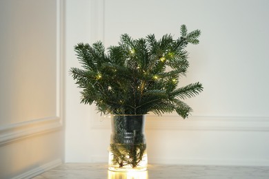 Photo of Beautiful fir tree branches decorated with Christmas lights on table near white wall