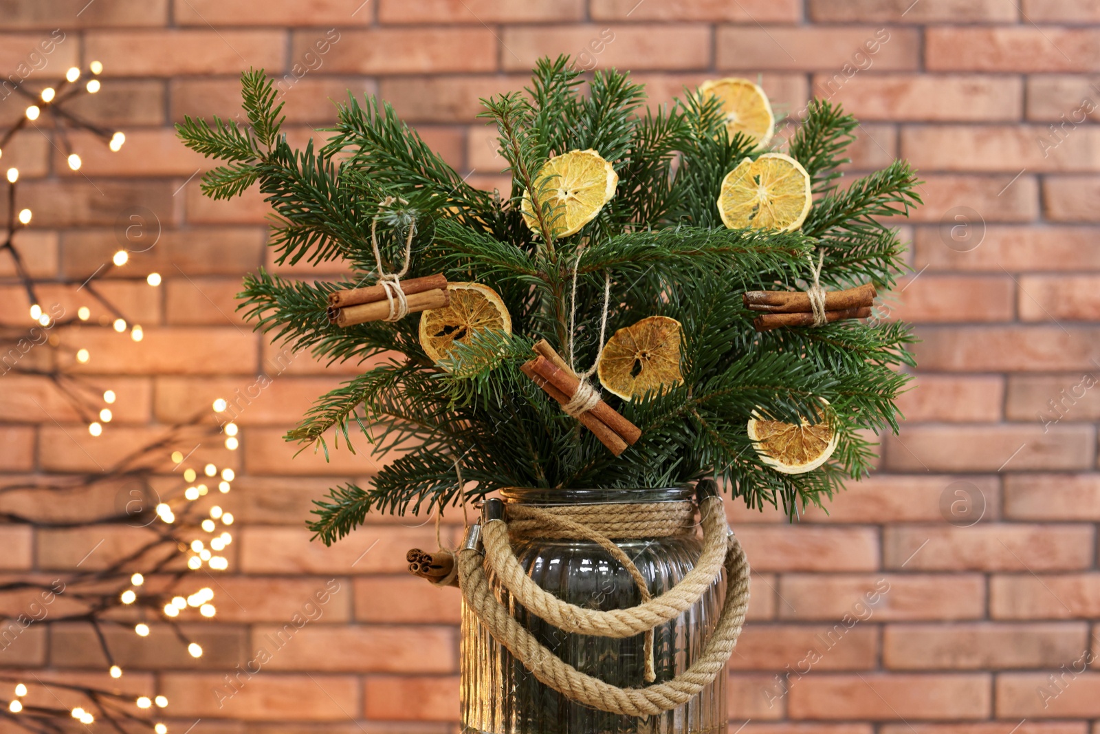 Photo of Beautiful fir tree branches decorated with dried orange slices and cinnamon sticks in vase against Christmas lights