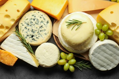 Photo of Different types of cheese, rosemary and grapes on dark textured table, flat lay