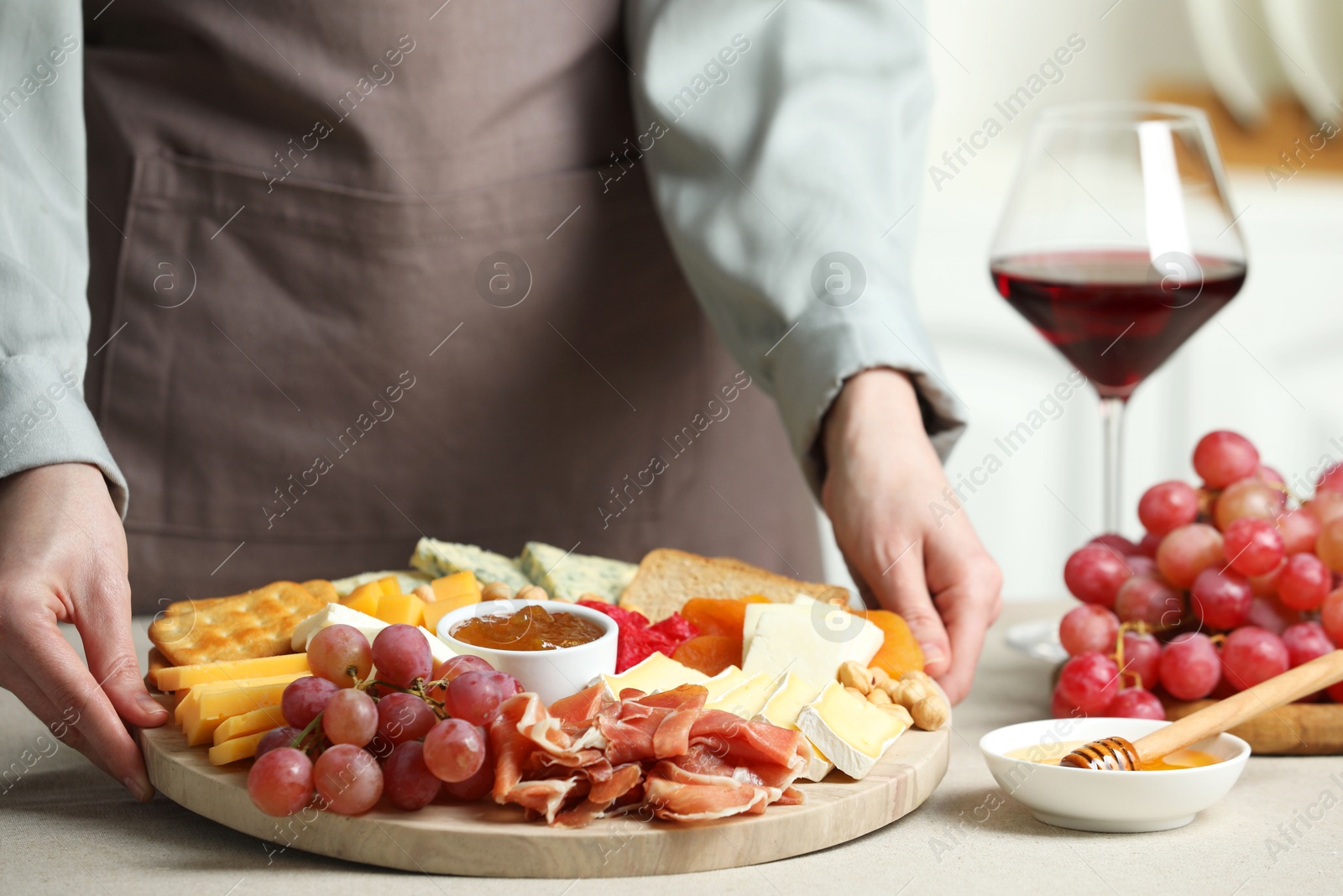 Photo of Woman holding board with different types of cut cheese and other snacks at light textured table, closeup