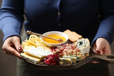 Photo of Woman holding board with different types of delicious cheese and other snacks on gray background, closeup