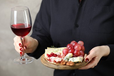 Photo of Woman holding board with different types of delicious cheese, other snacks and wine on gray background, closeup