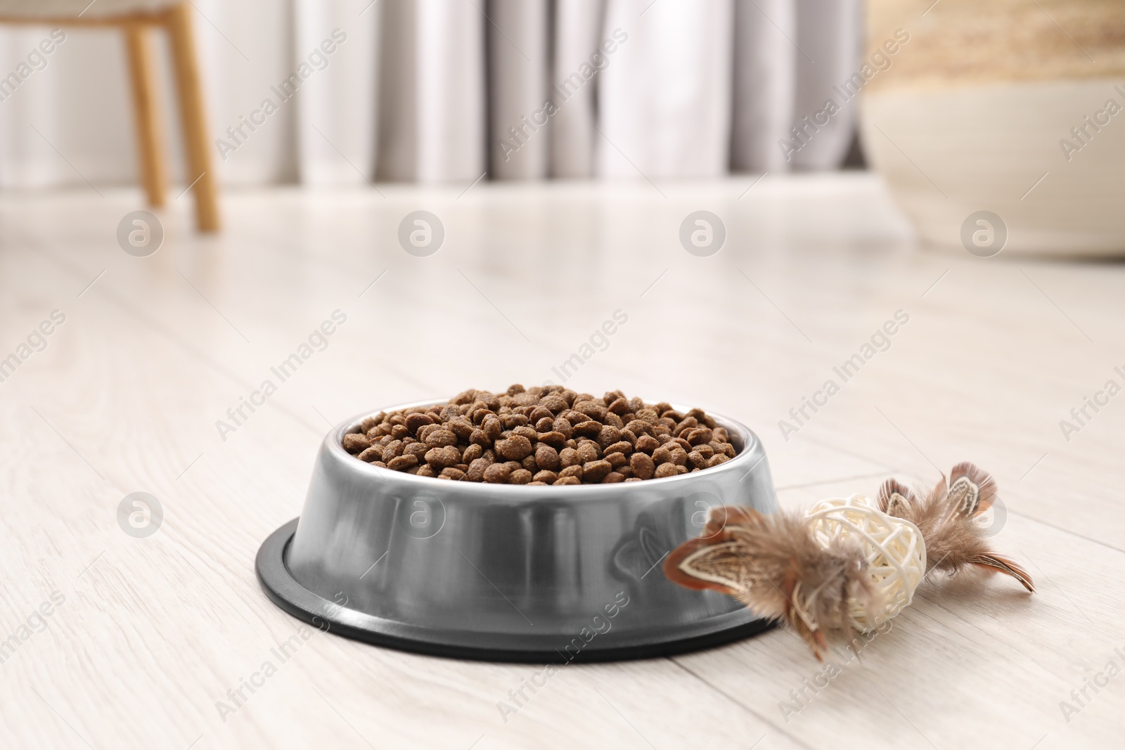 Photo of Dry pet food granules in feeding bowl and cat toy on floor indoors