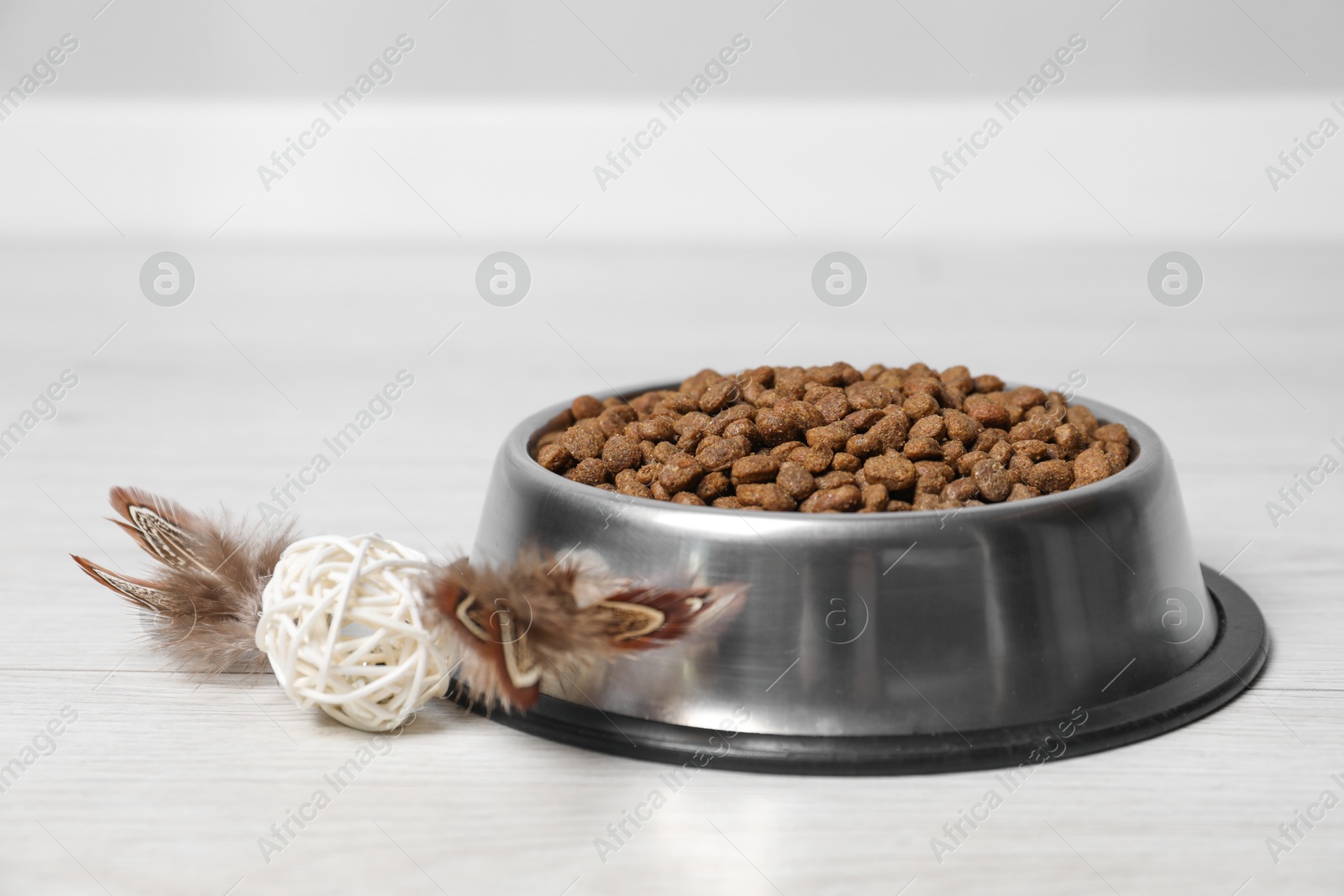 Photo of Dry pet food granules in feeding bowl and cat toy on floor indoors