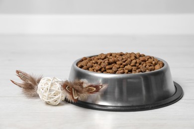 Photo of Dry pet food granules in feeding bowl and cat toy on floor indoors