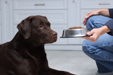 Photo of Woman giving bowl with dry pet food to her dog indoors, closeup