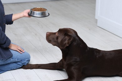 Photo of Woman giving bowl with dry pet food to her dog indoors, closeup