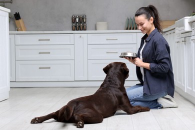 Photo of Woman with feeding bowl giving dry pet food to her dog indoors