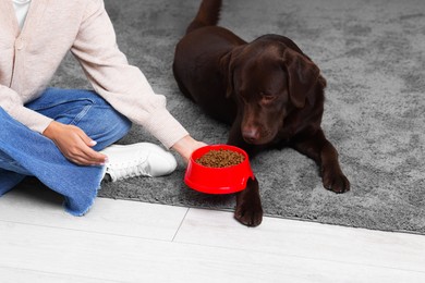 Photo of Woman giving bowl with dry pet food to her dog indoors, closeup