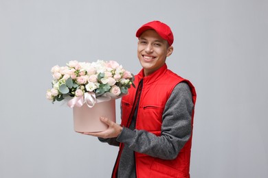 Photo of Smiling delivery man holding gift box with beautiful floral composition on grey background