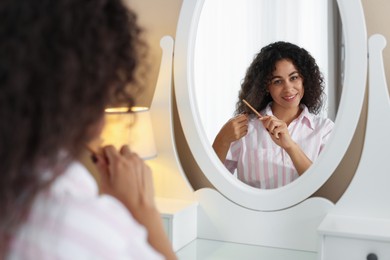 Photo of Smiling young woman brushing her curly hair with comb near mirror at home, selective focus