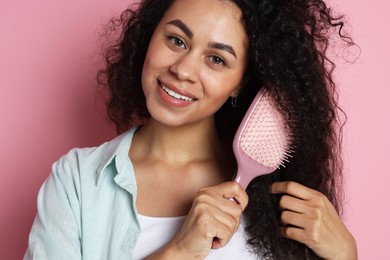 Photo of Smiling young woman brushing her curly hair on pink background