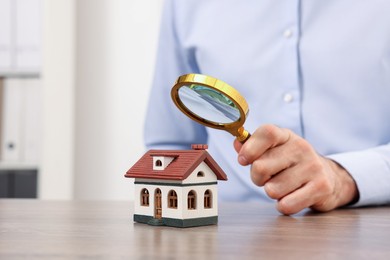 Photo of Real estate agent looking at house figure through magnifying glass at wooden table in office, closeup with space for text. Home appraisal