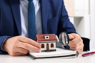 Photo of Real estate agent with house figure, notepad and magnifying glass at white table in office, closeup. Home appraisal