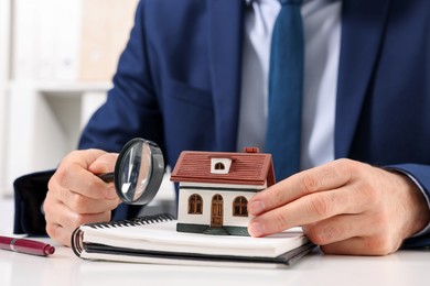 Photo of Real estate agent with house figure, notepad and magnifying glass at white table in office, closeup. Home appraisal