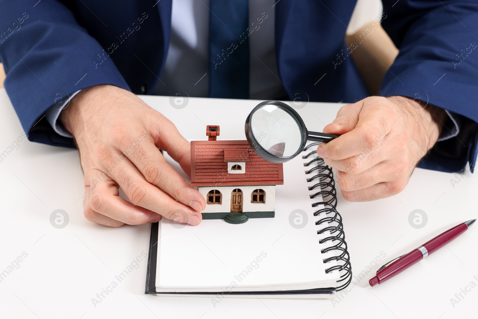 Photo of Real estate agent with house figure, notepad and magnifying glass at white table, closeup. Home appraisal