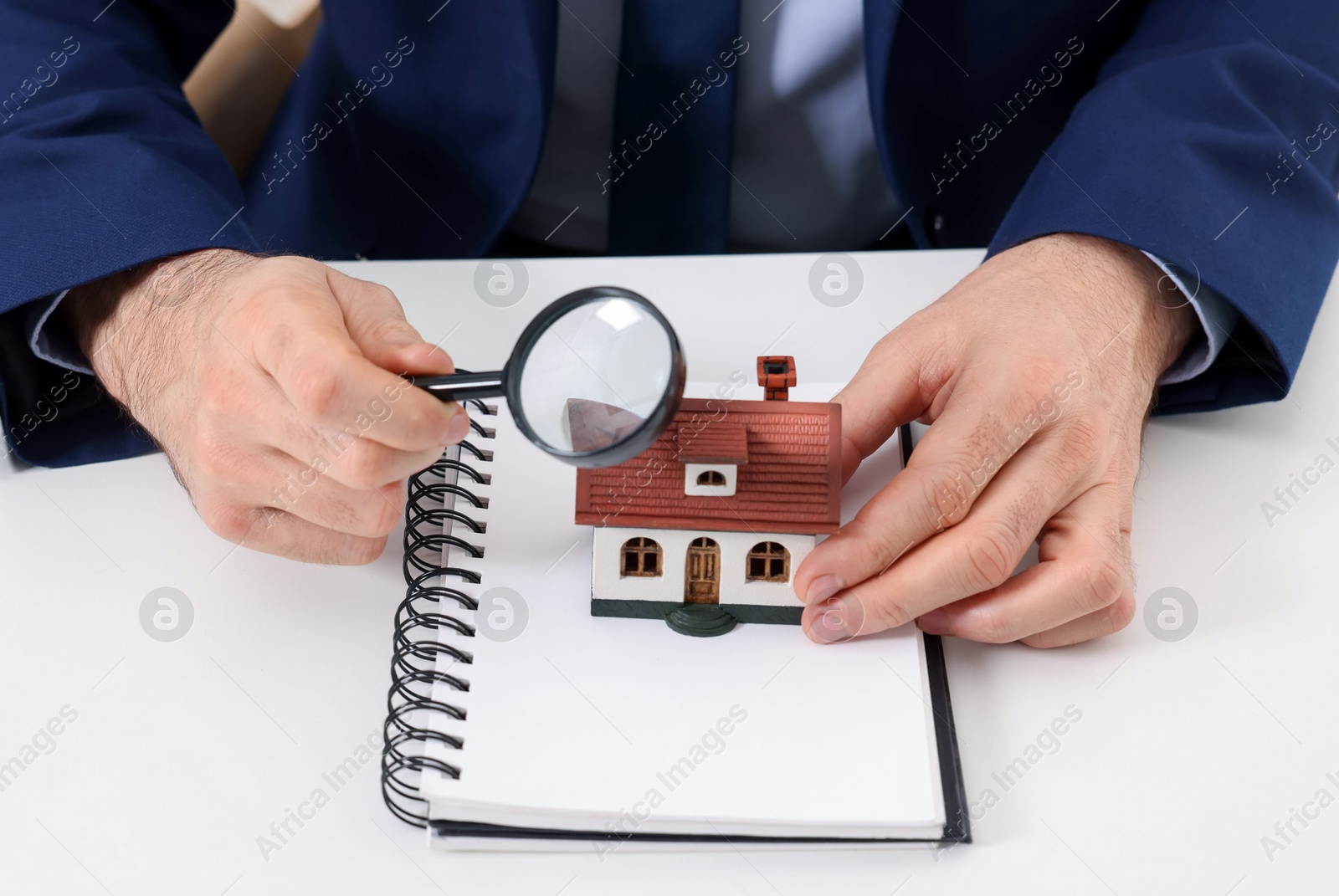 Photo of Real estate agent with house figure, notepad and magnifying glass at white table, closeup. Home appraisal