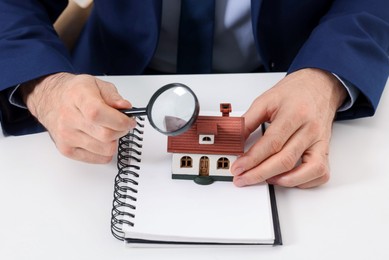 Photo of Real estate agent with house figure, notepad and magnifying glass at white table, closeup. Home appraisal