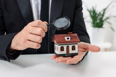 Photo of Real estate agent looking at house figure through magnifying glass at white table in office, closeup. Home appraisal